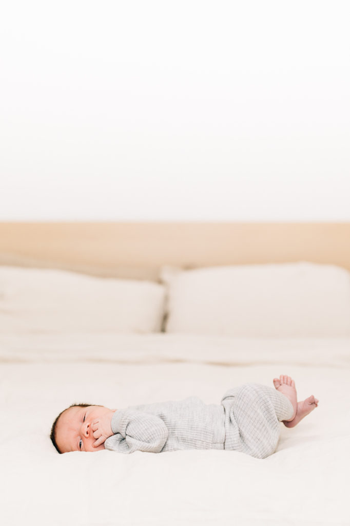 Family photographer in SLC Kailee Matsumura Photography captures a newborn laying on a bed eating his fingers. newborn baby #KaileeMatsumuraPhotography #SLCphotographers #motherhood #newbornphotography #Utahfamilyphotography #babyportraits