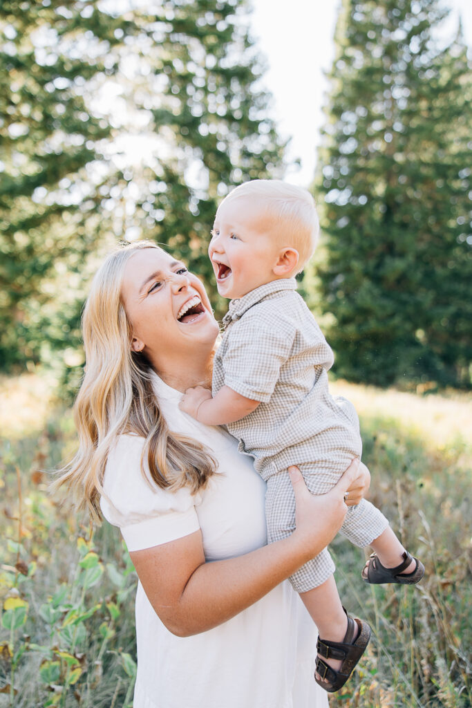 Mother and son spinning and laughing in a field with large trees and the sun. Toddler boy, mom with two year old boy. Mom spinning with little boy. Mom laughing with her son. Mom in white dress. Family outfit inspiration. 