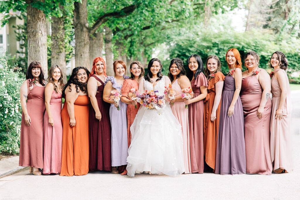 A group of bridesmaid in oranges, pinks, and purples stand under green leaf trees in the TN summer by Kailee Matsumura Photography. colorful bridesmaid dress summer wedding #KaileeMatsumuraPhotography #Bartlett,TN #MemphisPhotography #KaileeMastumuraWeddings #TNweddingphotography #summerwedding #colorfulwedding