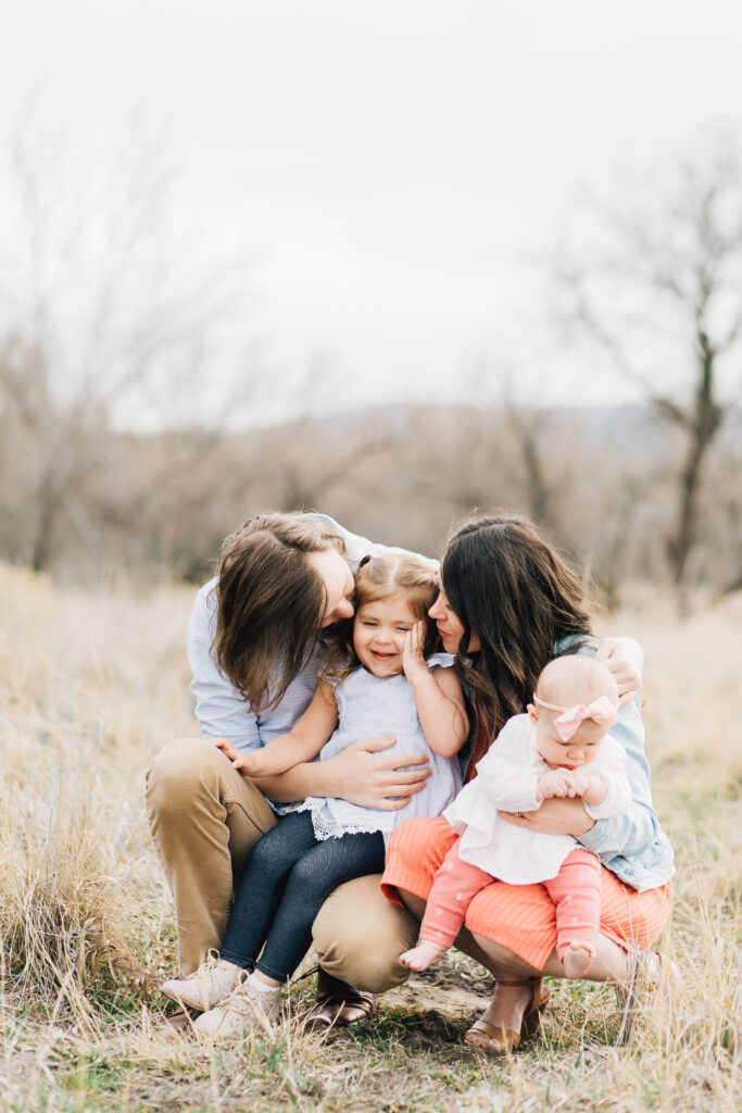 A very sweet, young family of four with two young daughters crouch low together. A mom and dad kiss their young daughters' cheeks. Blue and coral outfit inspiration
#KaileeMatsumuraPhotography #MeaningfulFamilyPhotoSessions #TennesseeFamilyPhotographer
