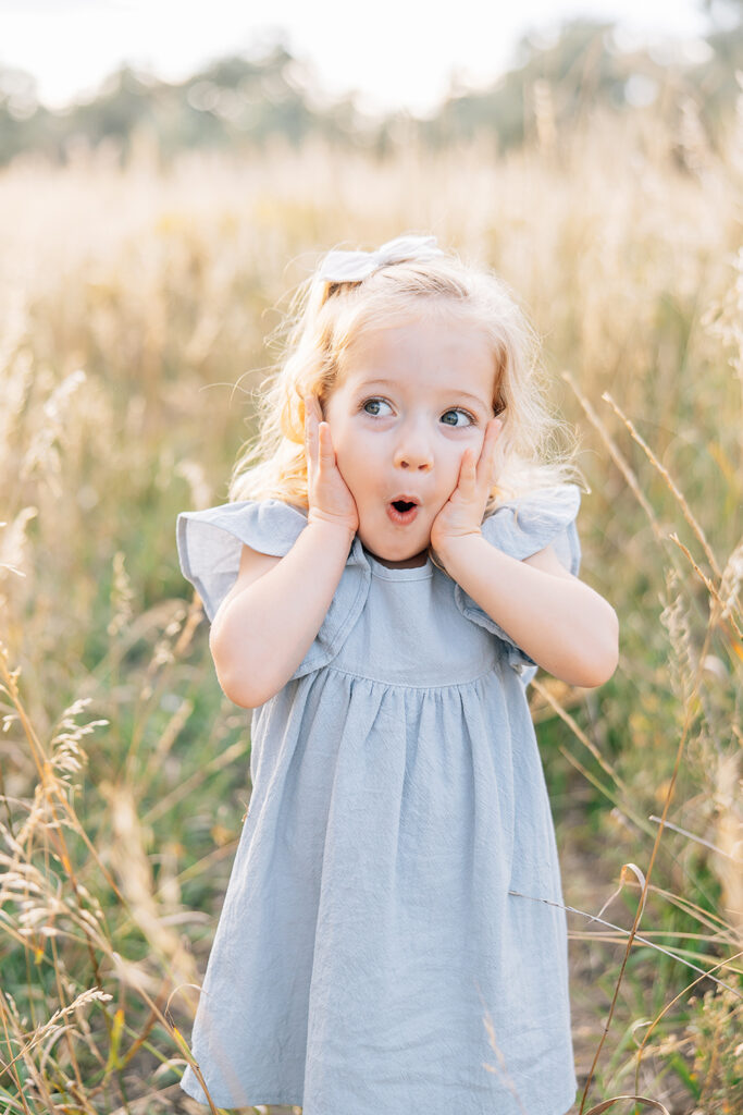 Documenting who you are is the goal for any family photographer; this adorable little girl shows her cute personality in a sunlit field with blonde curly hair and light blue linen dress. Funny faces kids family everyday stress-free #outdoorfamilyphotography #Tennessefamilyphotographer #mississippifamilyphotographer #tipsforsummerfamilyphotos
