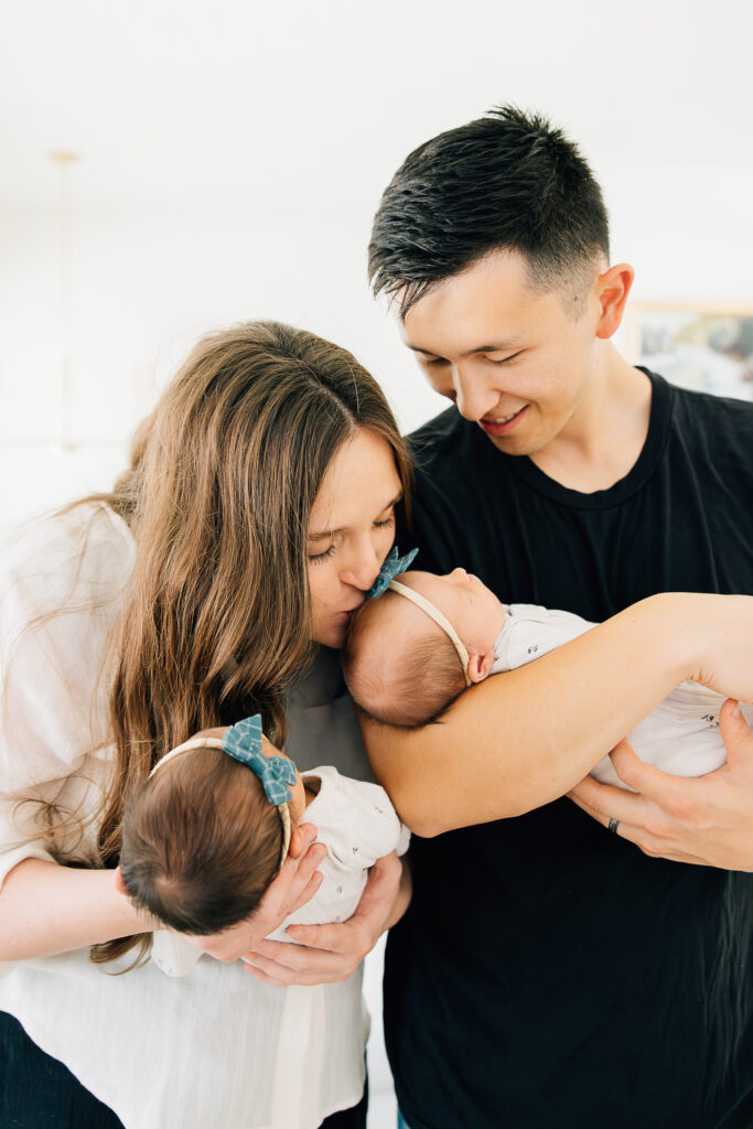 A mom and dad pose for their in home newborn session. mom kisses one of her twin girls on the head as she holds her other daughter. Neutral black and white casual, in home newborn photos by Memphis Newborn Photographer, Kailee Matsumura. 