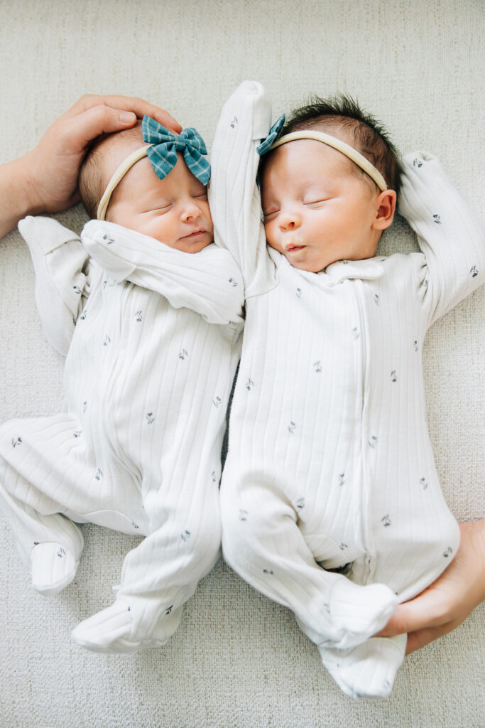 Photo of twin girls with their mom and dads hands to depict how small they are. They are wearing white ribbed pajamas with a simple floral pattern that is very minimalistic. 