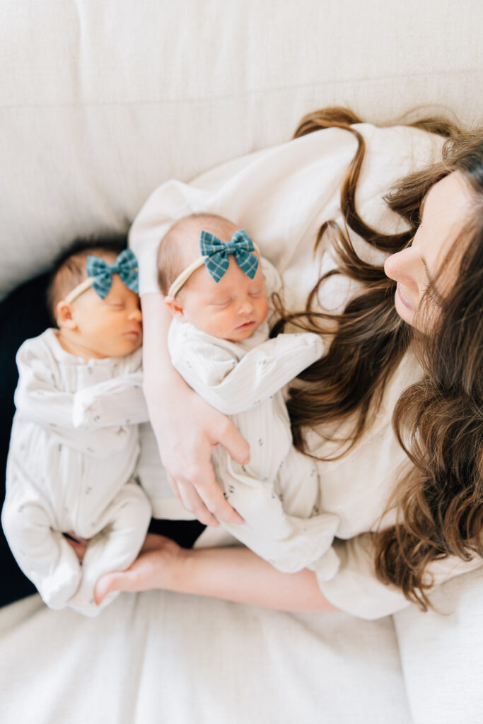 Mom holds her twin newborns on the couch during her Photoshoot. White and Blue Newborn session. Studio Newborn Session. In home lifestyle newborn session. Photos taken by Kailee Matsumura- Memphis Newborn Photography.