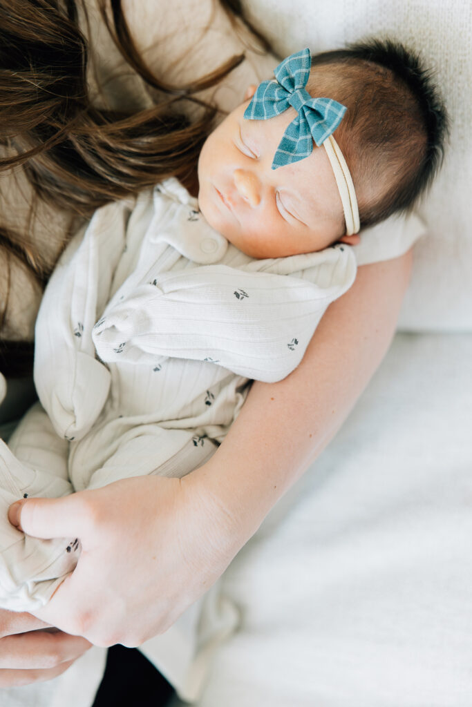 Newborn in white and blue sleeping in moms arms. Newborn girl has lots of hair, all dark. Photoshoot was in their home. 