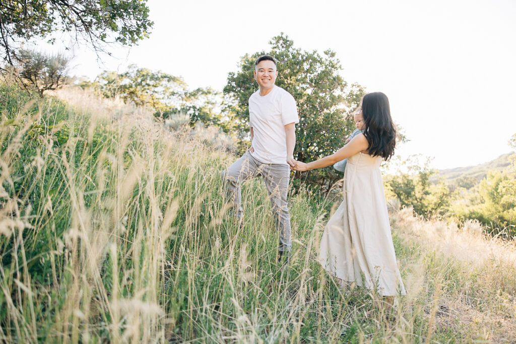 Dad smiles at camera while leading mom and newborn baby up a hill. Summer outfit inspo. Photos with long green grass in Germantown, TN. Taken by Kailee Matsumura, a Germantown Newborn Photographer. 